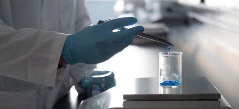 Person in a white lab coat and blue glove scoops blue powder into a beaker. 