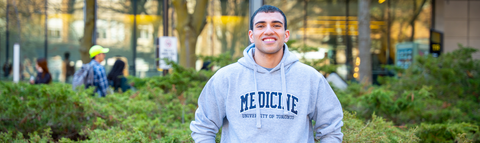 Ahmed standing outdoors in the rock garden at Waterloo's campus, wearing a grey hoodie with the text “Medicine, University of Toronto.” Trees, shrubs, and a glass building are visible in the background.