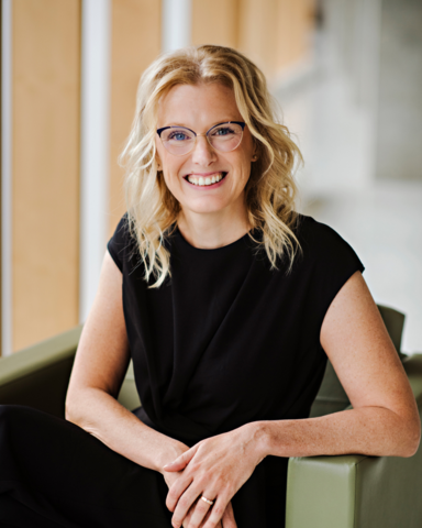 A headshot of Andrea. She is wearing a black t-shirt and pants and is sitting on a chair with her hands crossed. 