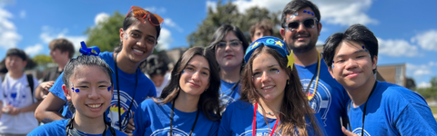 Seven students wearing blue t-shirts and smiling at an outdoor orientation event.