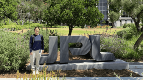 Ariana standing beside a large gray "JPL" sign outdoors at NASA’s Jet Propulsion Laboratory.