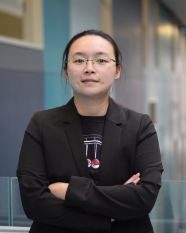 A headshot of Brenda. She is wearing a black blazer and t-shirt. She is standing with her arms crossed. 