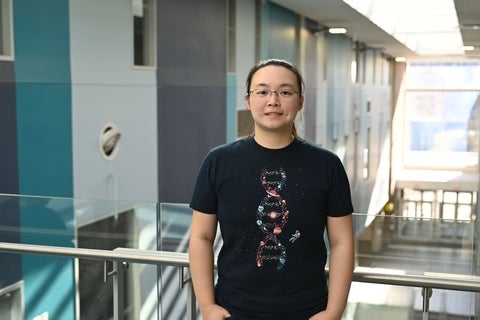 Brenda Lee is wearing a black t-shirt, her brown hair is in a ponytail, and she is wearing glasses. She is standing in a large hallway with blue squares painted on the wall.