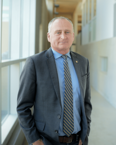 Dean Chris Houser standing in a long hallway with his hands in his pockets. He is wearing a blue dress shirt, a grey tie, and a grey blazer.