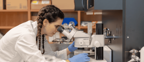 A science student wearing a white lab coat looking into a microscope in a lab