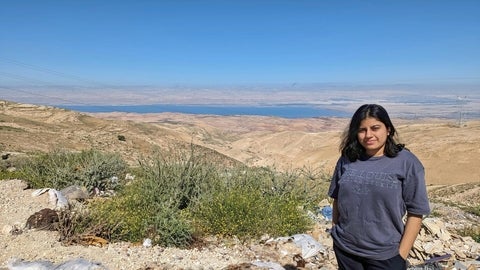 Aparajita Bhattacharya, a graduate student standing in an open area with sand dunes, plants, and a source of water in the background