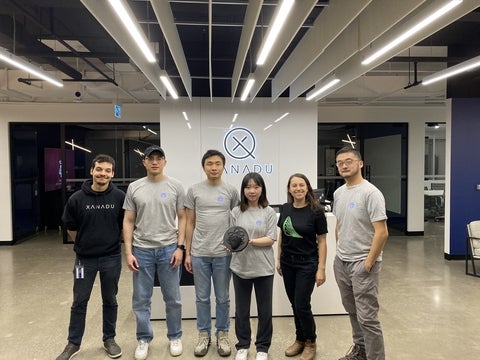 A group of six stand in an open concept office. A white back drop has a lit up logo that says "Xanadu."