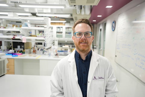 Professor Dale Martin is standing in the NeurdyPhagy research lab. He is wearing a white lab coat, a blue collared shirt, and blue glasses.