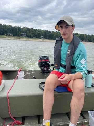 Joey Ruck wearing a life jacket, a turquoise t-shirt, and a ball cap. He is sitting at the back of a boat and steering it. 
