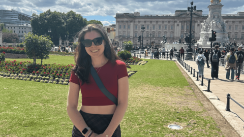 Emma Ross smiling outdoors in front of Buckingham Palace in London, wearing sunglasses, a red top, and a crossbody bag, with gardens and crowds of people in the background.