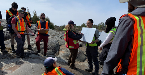 Jen Parks surrounded by a group of students wearing an orange safety vest. She is delivering a lesson outside at a waterfront. She is writing an explanation on a small whiteboard.