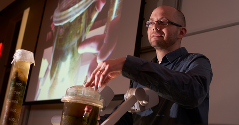Josh Neufeld standing at the front of a classroom delivering a lecture. The room is dark and he is projecting an experiment on the projector screen behind him. 