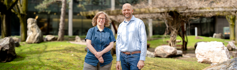 Jen Parks is on the left and is wearing a dark blue button up t-shirt. Josh Neufeld is on the right wearing a light blue button up long sleeve. They are standing in the rock garden at the University of Waterloo.