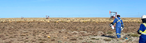 Gabriella walking on soil with oil wells in a flat, dry field. 