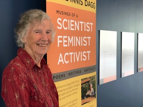 Anne is wearing a red shirt and is standing next to a sign that says "Scientist, feminist, activist."