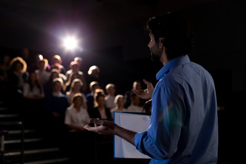 A male student stands in a dark auditorium full of people with a stage light shining on him. He is speaking to the audience. 