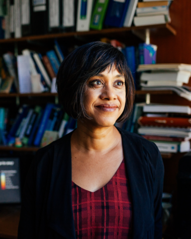 A headshot of Nandita. She is wearing a read striped shirt and a black blazer. There are book shelves in the background. 