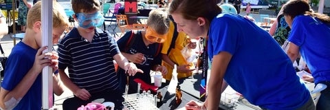 A Waterloo Science outreach volunteer supervises while a child runs an experiment at an outdoor event.