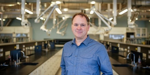 Dr. Pierre-Nicholas Roy is wearing a blue shirt and standing in a lab. 