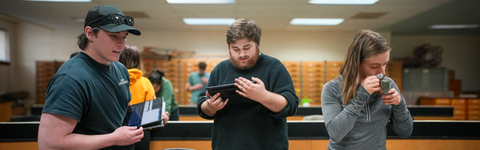 Three people stand at lab benches in a classroom-style science space, each examining a handheld device or tablet, with wooden cabinets and additional people working in the background under fluorescent lighting.
