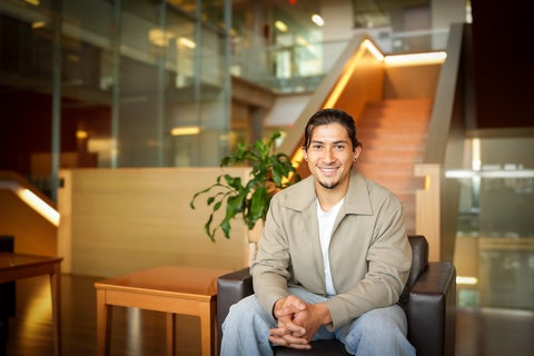 Kent Ueno is wearing a beige coat and white t-shirt and he is sitting on a brown leather chair with glass windows in the background.