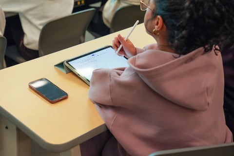A student writing notes on an ipad at a desk.