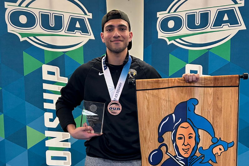 Ahmed holding an OUA medal and a clear glass award plaque in front of blue and green banners reading “OUA” and “Championship.”