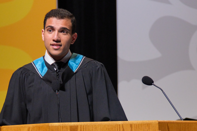 Ahmed wearing a graduation gown standing behind a wooden podium, speaking into a microphone on a stage.