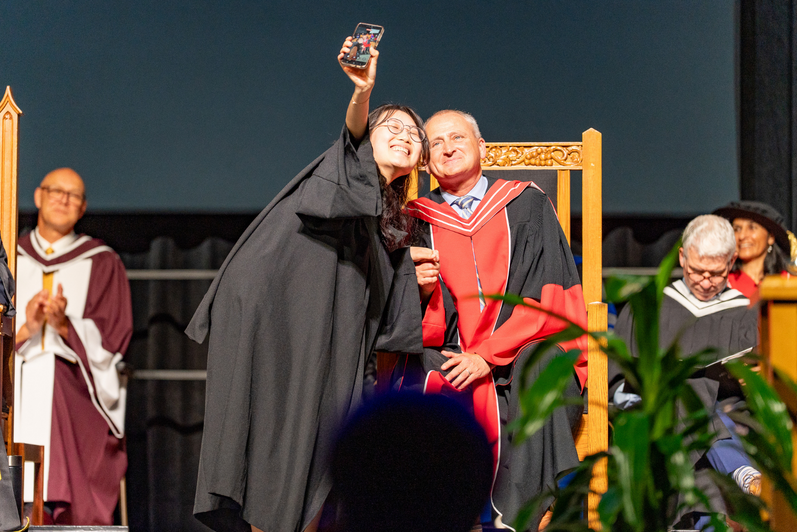 Dean Chris Houser on stage at convocation taking a selfie with a student. 