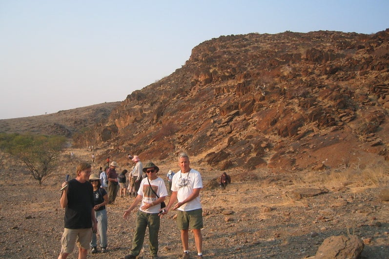 Shaun Frape holding a rock hammer, standing with several people at a dessert field site. A large red rock outcropping is behind them
