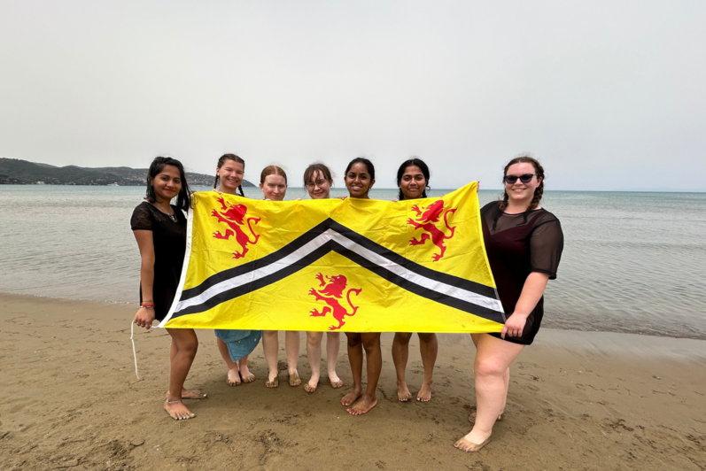 Seven students holding the University of Waterloo flag on a beach in Italy on a cloudy day. 