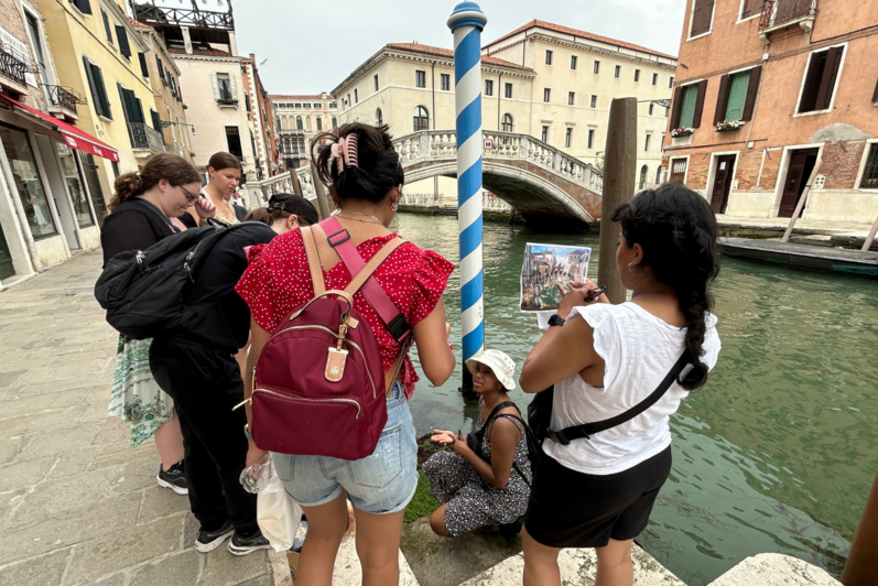 Two students looking out at a canal in Venice. They are holding up an older image of Venice to compare the water levels. 