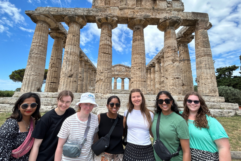 Seven students standing in front on ancient ruins in Italy. 