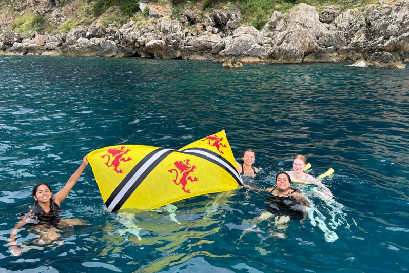 Four students swimming near the island of Capri. The are holding up the University of Waterloo flag.