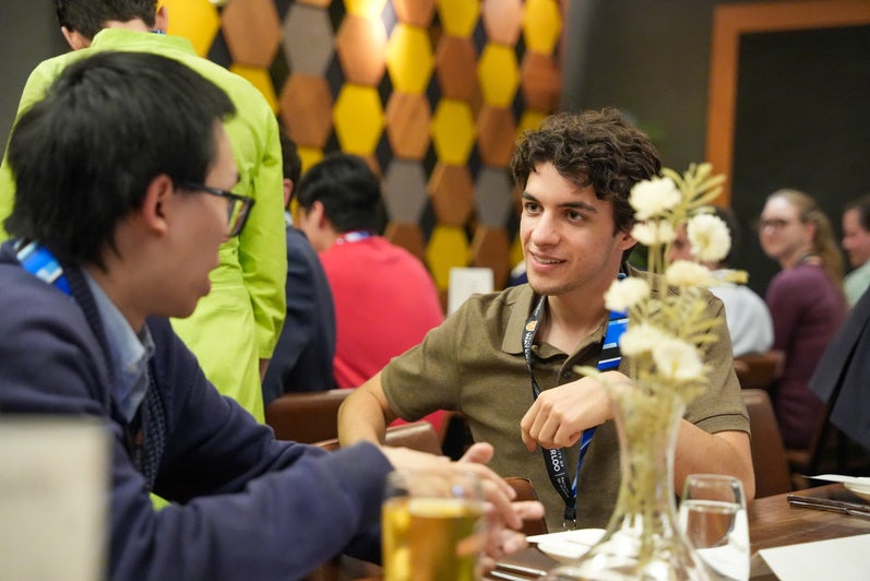 Two students talking at table with other people in the background. 