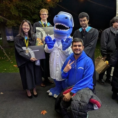Four graduates dressed in their convocation gowns and hoods posing with Cobalt the mascot