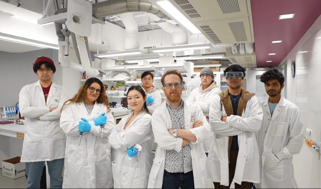 Eight scientists in the NeurdyPhagy lab. They are all posing with their arms crossed. The scientists are wearing white lab coats with blue gloves.