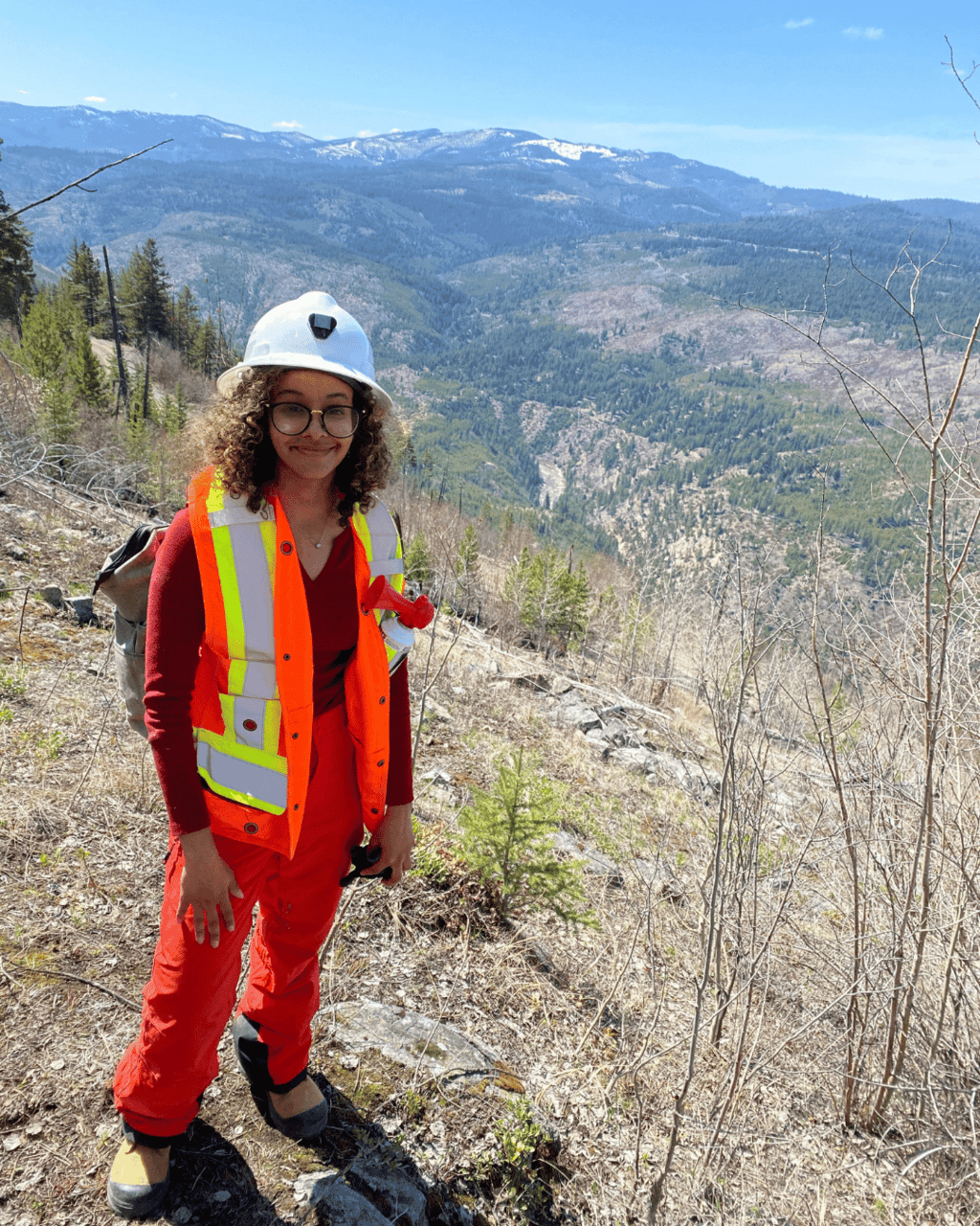 Olla is standing on a hillside with mountains in the background. She is wearing a white hard hat and an orange safety vest and pants. 