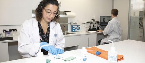 Female working in at a lab bench with male at fume hood in background