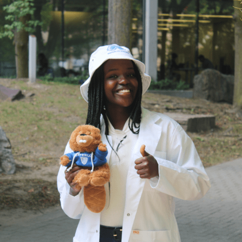 A woman smiling with a thumbs up. She is wearing a white bucket hat, a white lab coat, and his holding a stuffed animal.