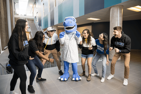 Blue dinosaur dressed in a white coat and safety goggle is surrounded by six happy students. 