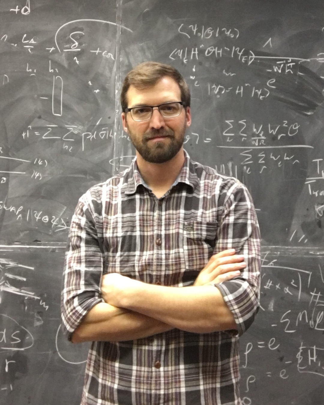 Roger Melko is a Professor of Physics in the Faculty of Science. He is standing with his arms crossed in front of a blackboard.