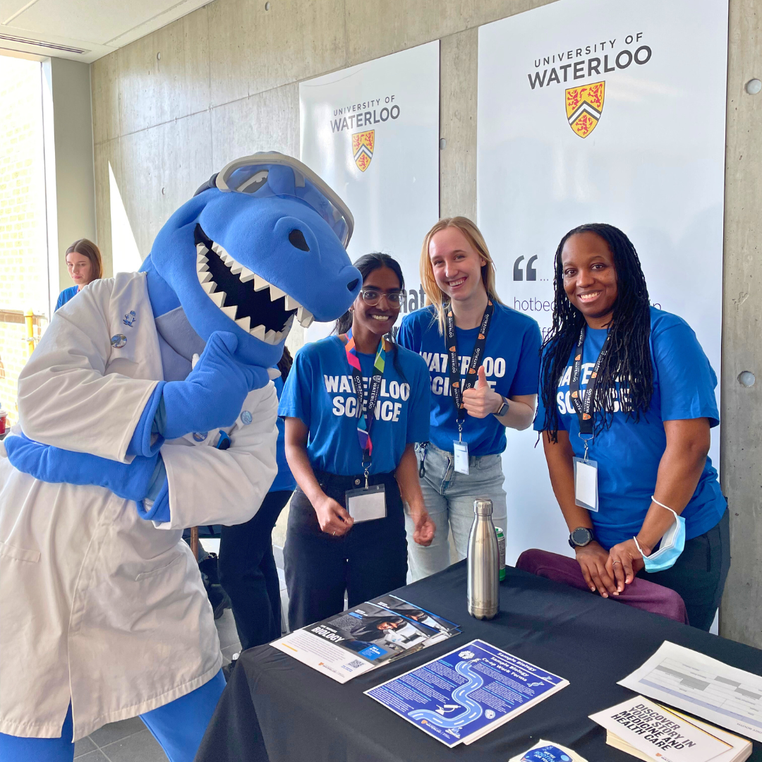 Three students wearing blue Science t-shirts in front of University of Waterloo banners. Cobalt the blue mascot dinosaur is posing next to them.
