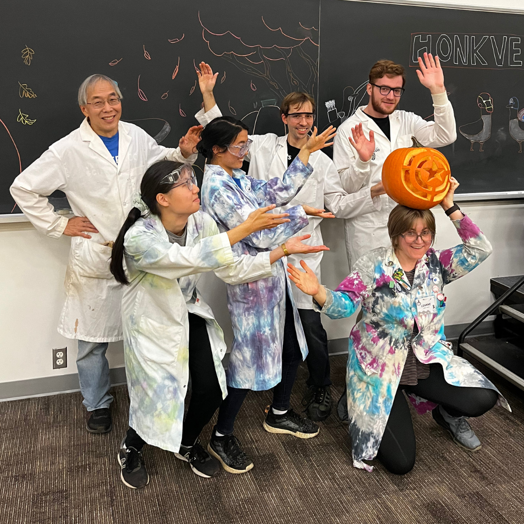 Six chemists pose in tie dye lab coats. One chemist is holding a pumpkin over her head. 