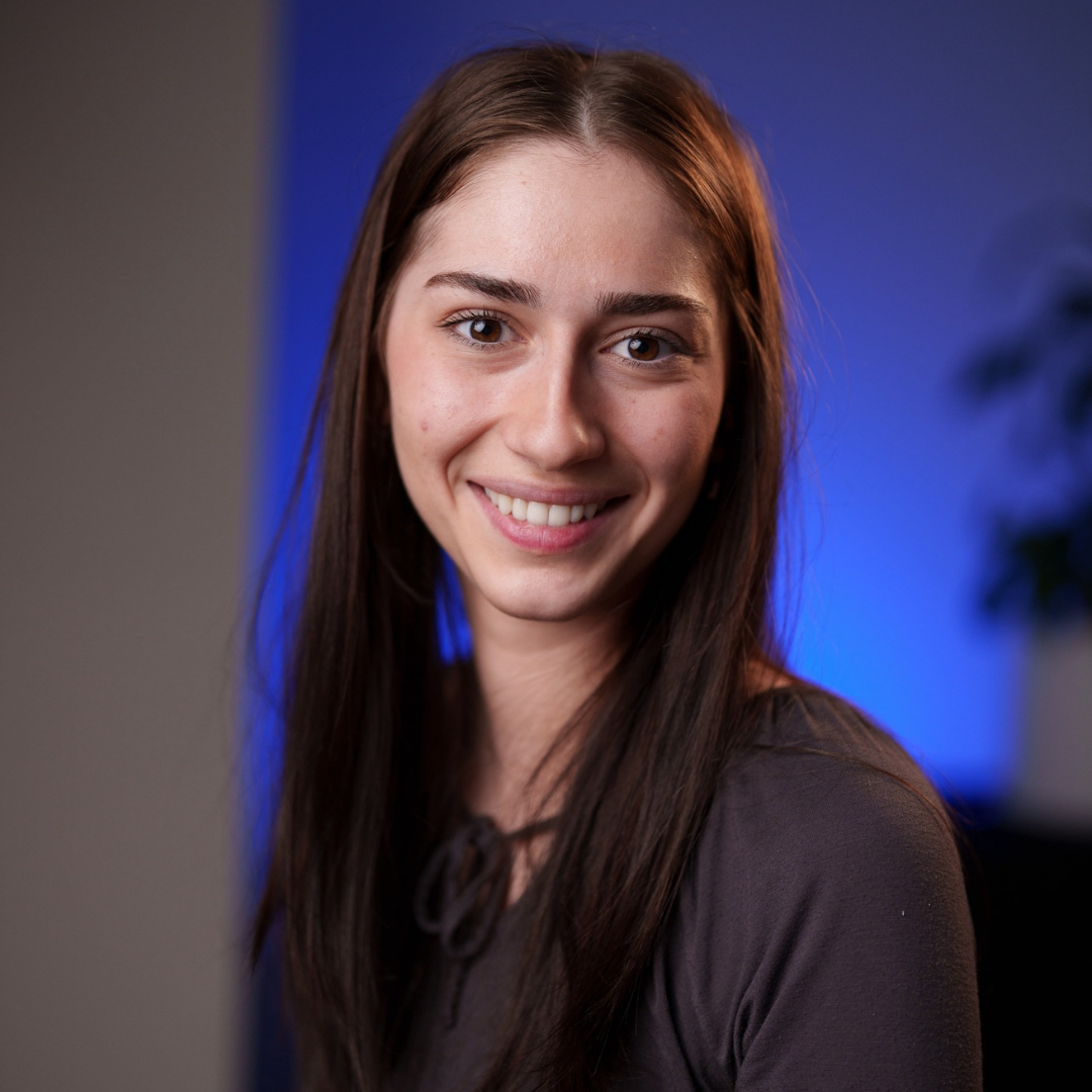 Milena Gojsevic has brown hair and brown eyes and is wearing a grey long sleeve shirt. She is against a blue lit background.