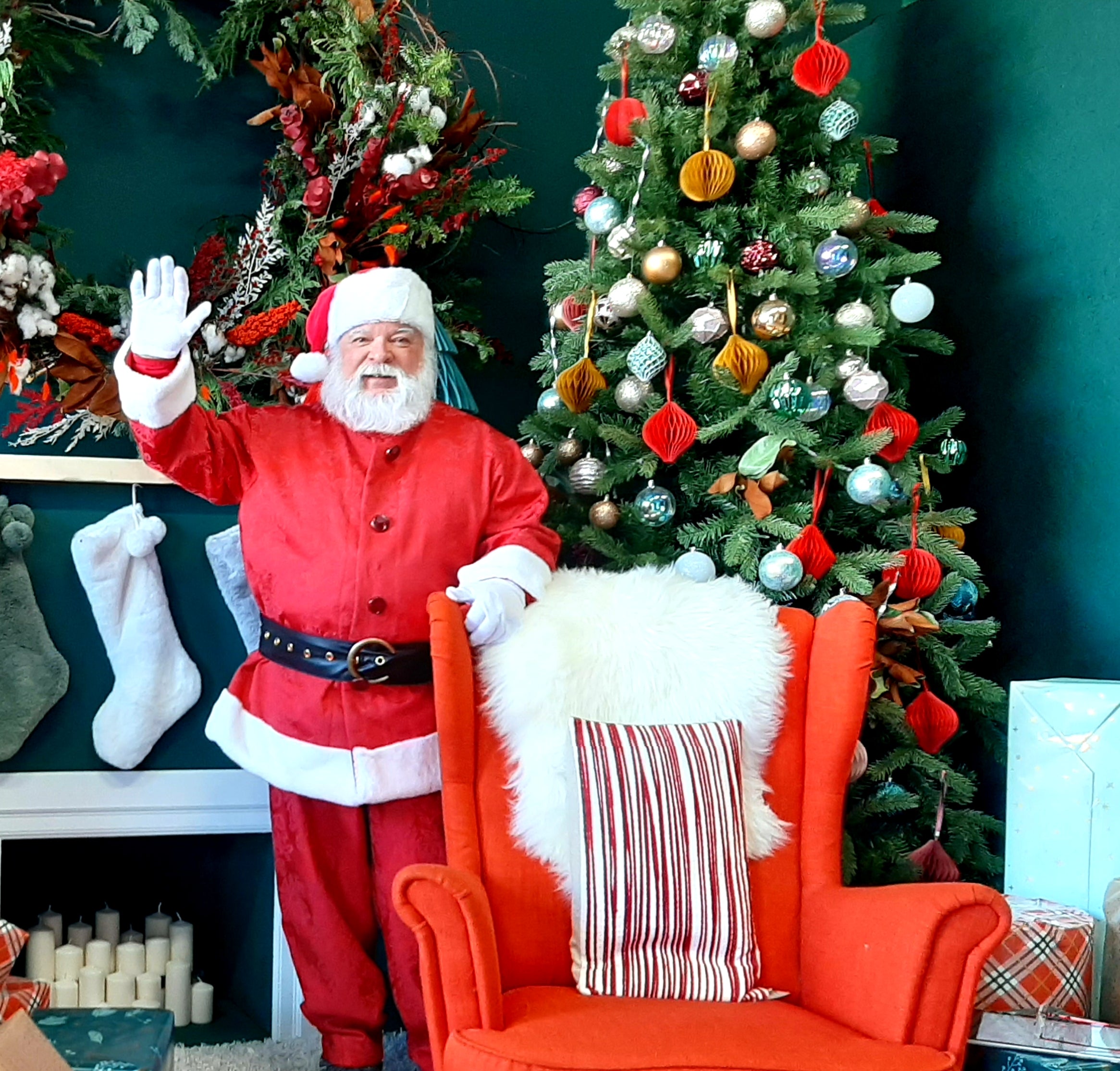 Santa standing in front of a decorated Christmas tree and fireplace, waving at the camera