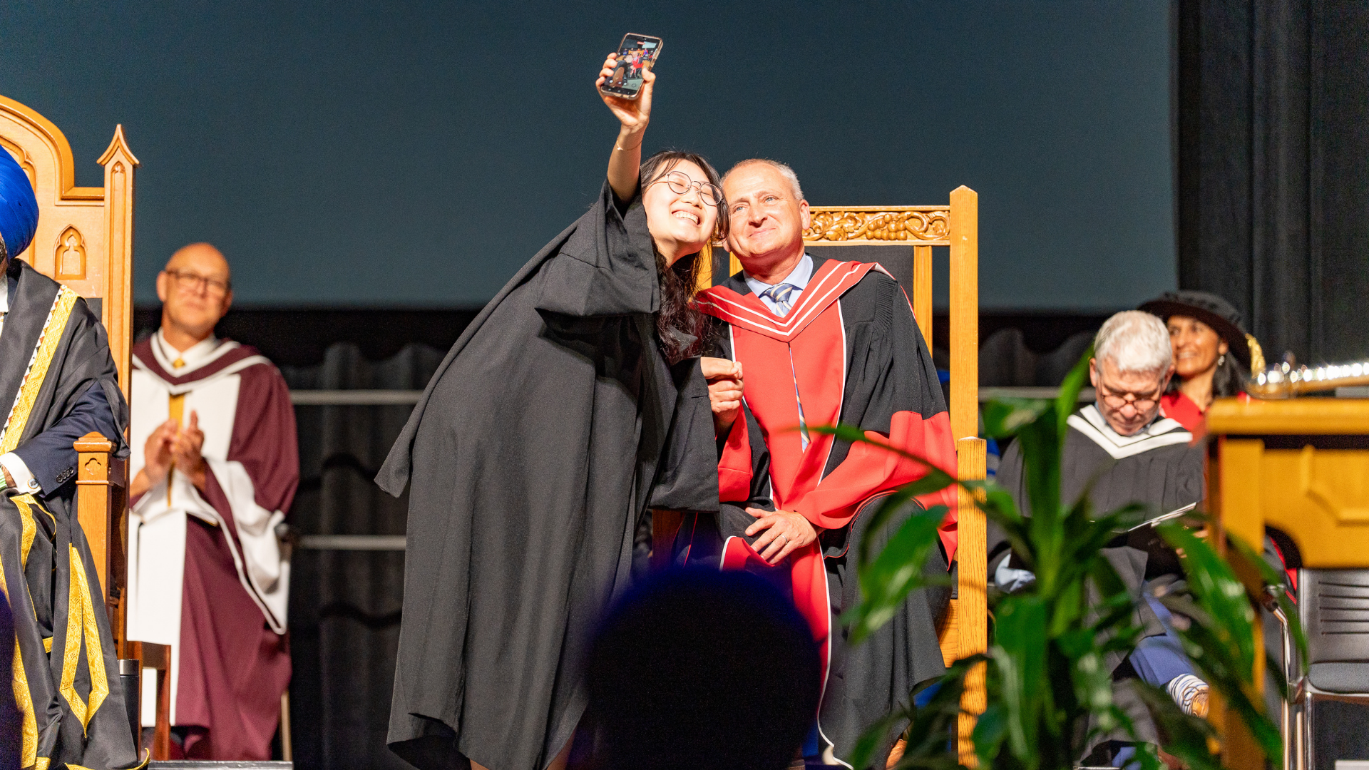 Dean Chris Houser on stage at convocation taking a selfie with a student. 