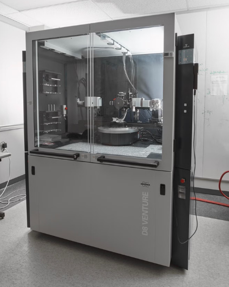A large instrument filling a room. There are clear glass doors on the front, which open out from the middle. Inside the cabinet is an intricate machine at the back of the cabinet, a track across the top of the cabinet, and a base below the intricate machine. The cabinet says Bruker D8 Venture.