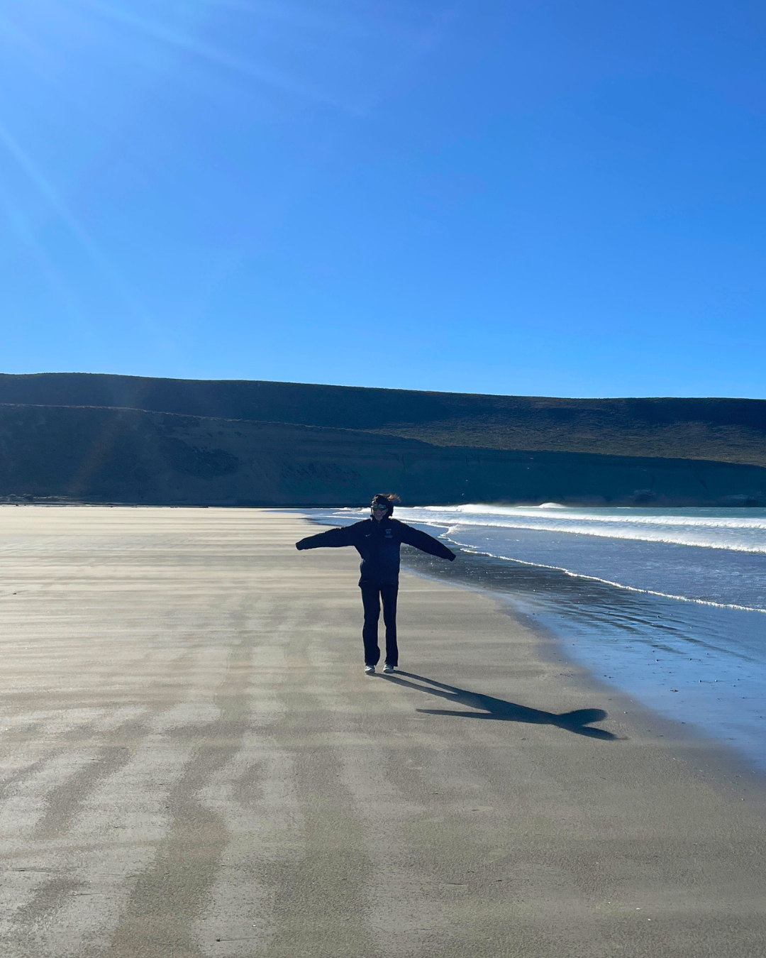 Gabriella standing on a beach on a sunny day, surrounded by sand, water and mountains.