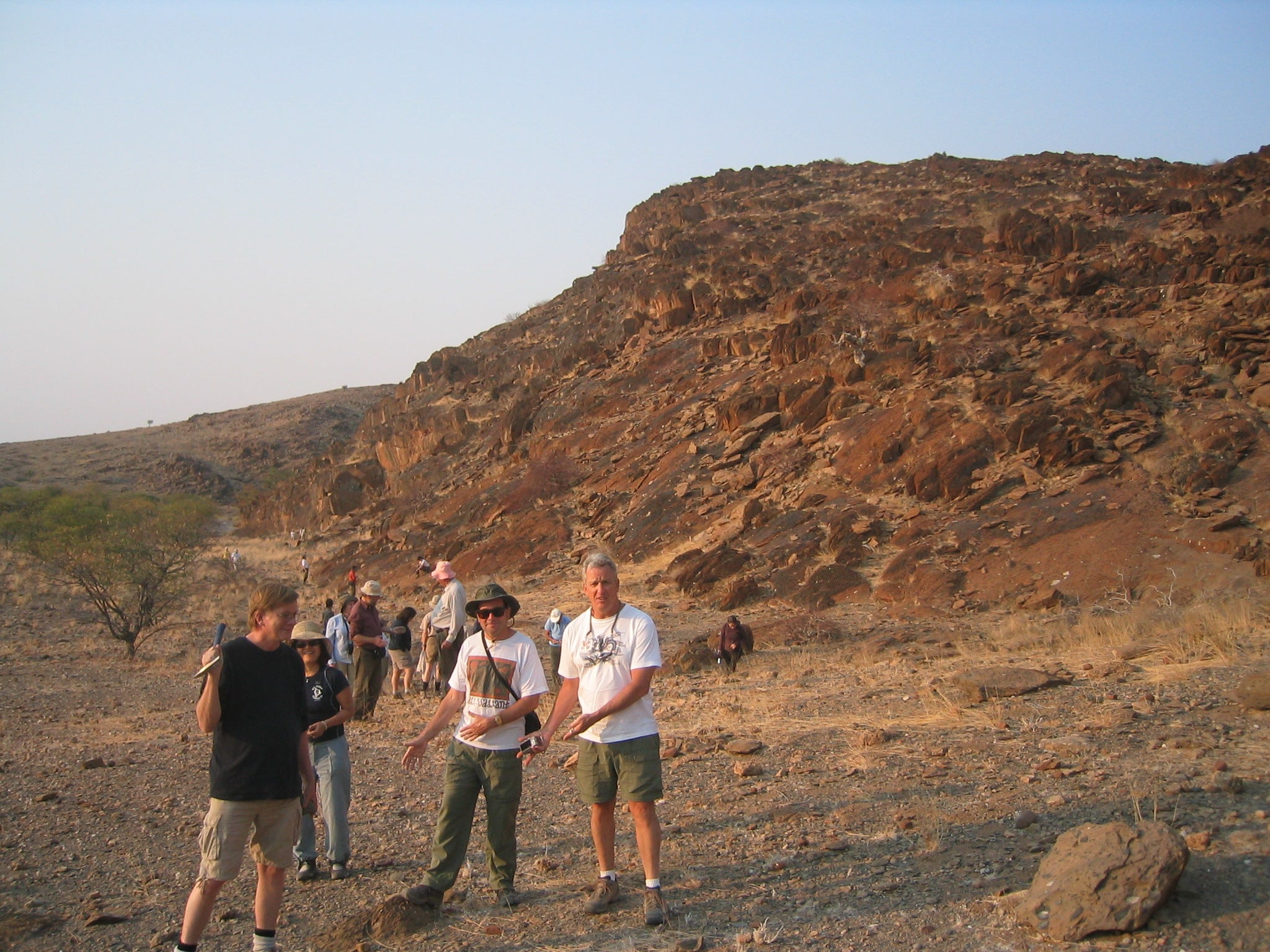 Shaun Frape holding a rock hammer, standing with several people at a dessert field site. A large red rock outcropping is behind them
